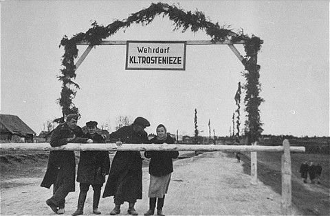 Three men and a woman stand behind the guard rail at the entrance to the Maly Trostinets concentration camp